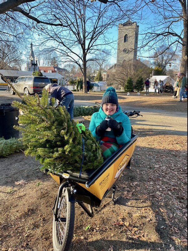 A young person bundled in winter clothes sits inside the cargo compartment of a bike with a Christmas tree.
