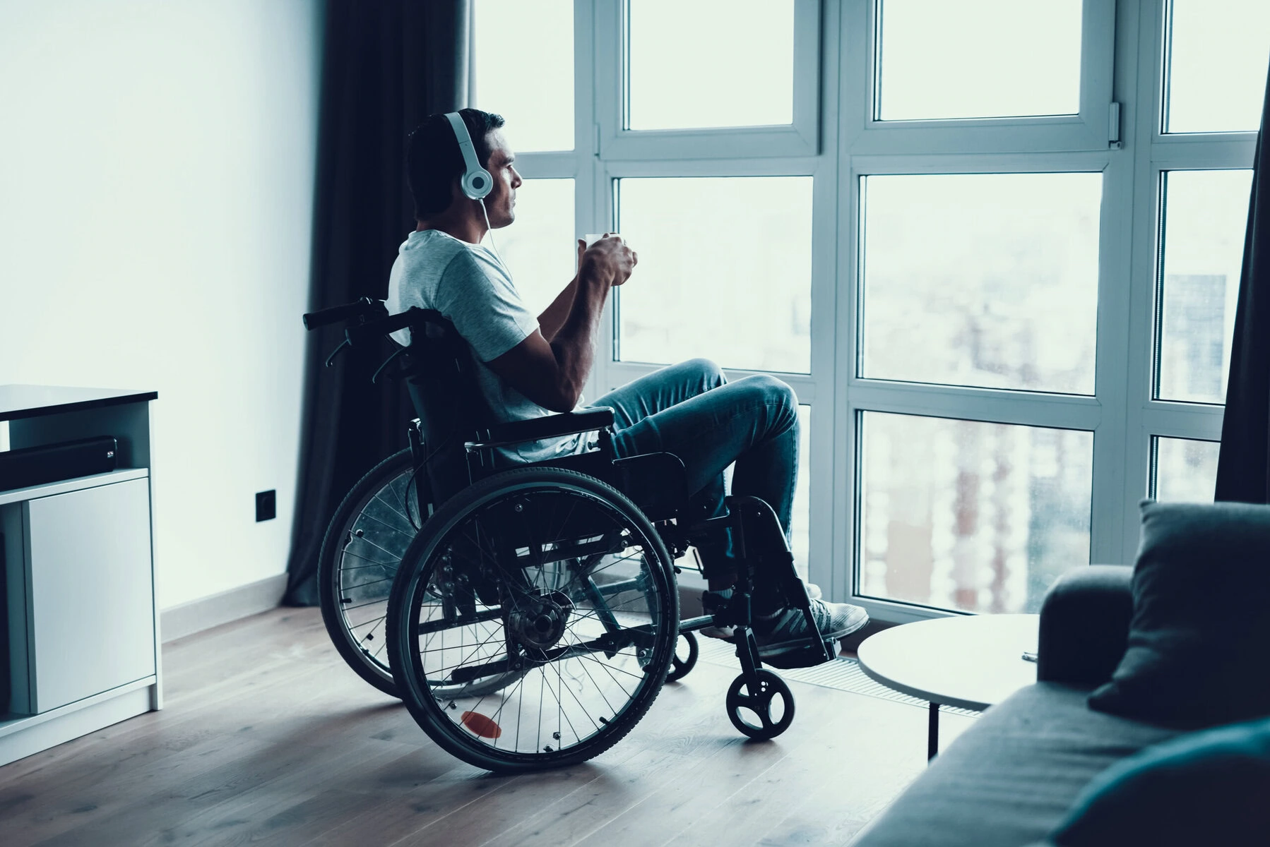 A man in a wheelchair looks out a window. He is wearing headphones.