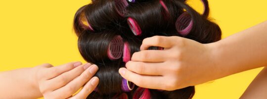 A woman putting curlers in her hair. A rear view with a yellow background.