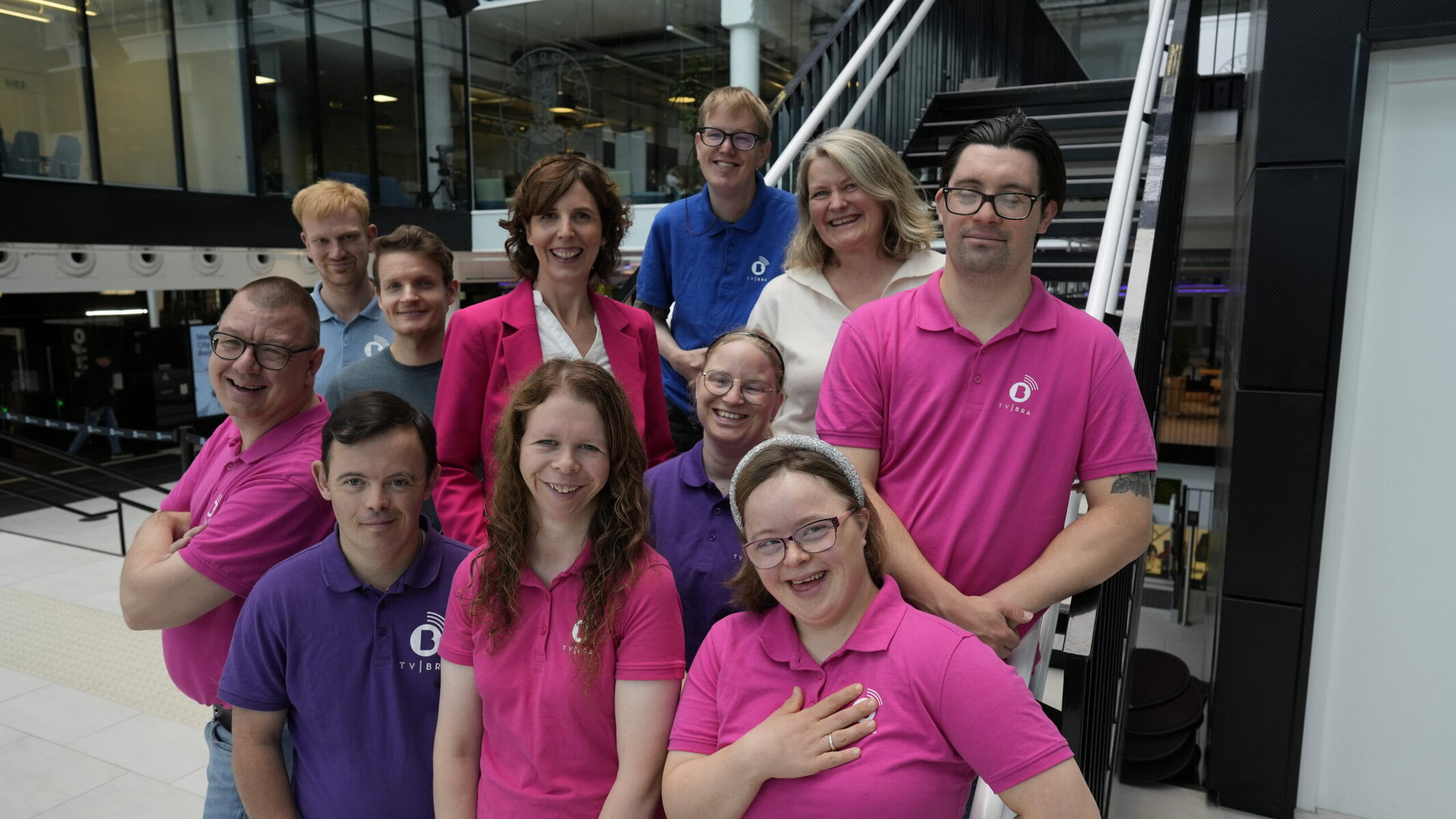 The TV Bra crew gathers at the foot of a staircase. They are wearing mostly pink and purple.